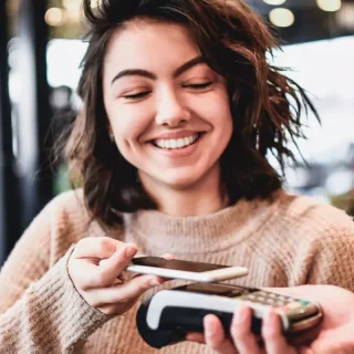 Eine lächelnde Frau benutzt ihr Smartphone, um in einem belebten Café an einem Kartenleser kontaktlos zu bezahlen.