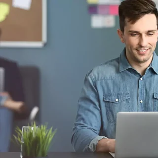 Ein Mann im Jeanshemd verwendet in einer geschäftigen Büroumgebung einen Laptop zur Marketingunterstützung, während im Hintergrund Kollegen arbeiten.